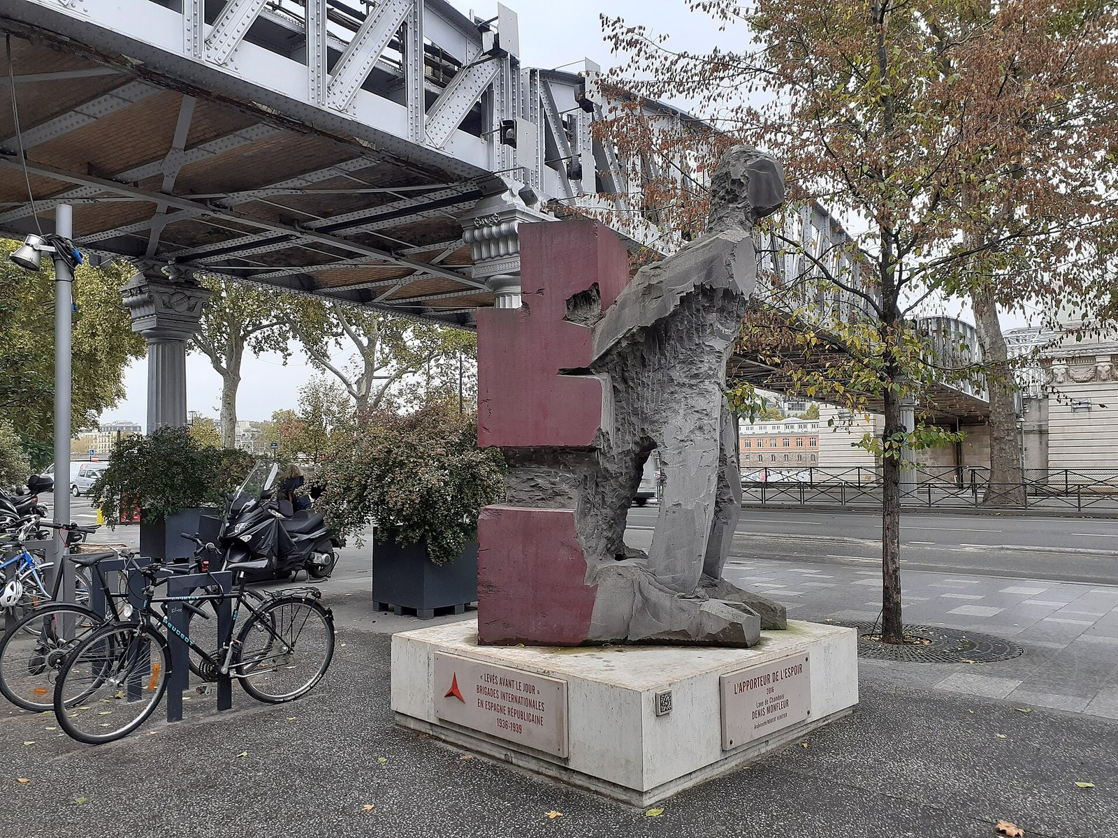 L’Apporteur de l’espoir de Denis Monfleur, hommage aux Brigades internationales, à la gare d’Austerlitz — Cahtls · CC BY-SA 4.0 — Wikimedia Commons Photographie : sous le viaduc du métro, une haute sculpture de pierre rouge aux formes taillées, sur un socle, près de vélos garés