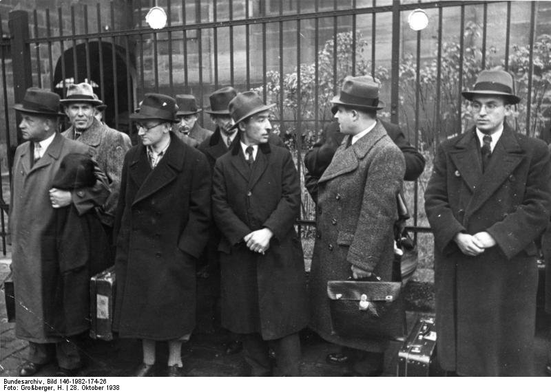 L’expulsion des Juifs polonais à Nuremberg, le 28 octobre 1938 — H. Großberger · Bundesarchiv, Bild 146-1982-174-26 · CC BY-SA 3.0 de — Wikimedia Commons Photographie en noir et blanc : des hommes en manteau et chapeau, valises à la main, attendent devant une grille