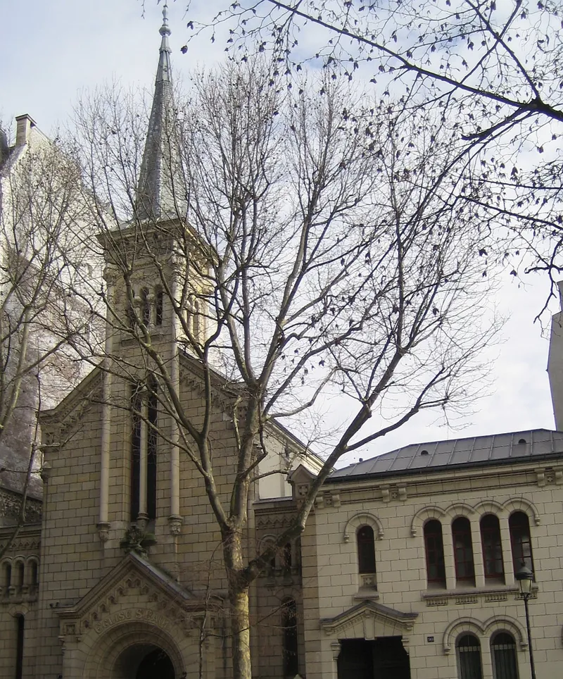 Photographie : la façade et le clocher pointu d'une église de pierre, derrière des arbres
