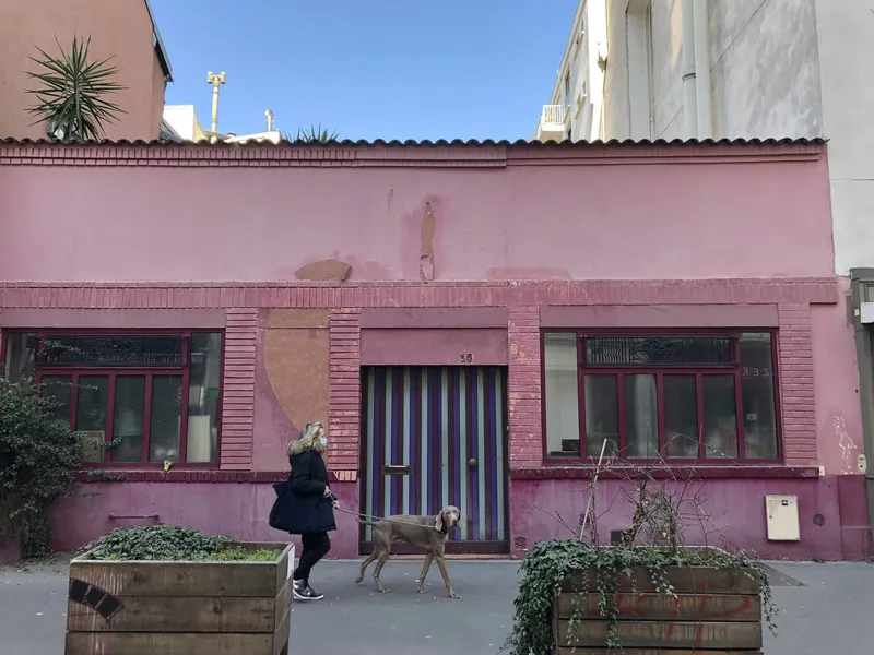 Photographie : une petite maison rose d'un étage sur la rue Daguerre, deux larges fenêtres encadrant une porte, des passants et un chien devant