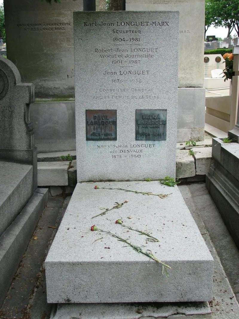 Photographie : une stèle de granit gravée de noms, avec deux plaques de bronze, au-dessus d’une dalle où l’on a déposé des œillets rouges