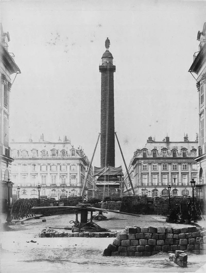 Photographie : la colonne Vendôme, retenue par des câbles, au centre de la place ; au premier plan, une barricade de pavés