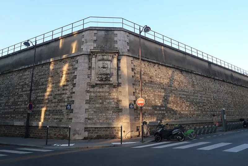 Photographie : un haut mur de soutènement en pierre meulière, surmonté d'une grille, au coin de deux rues