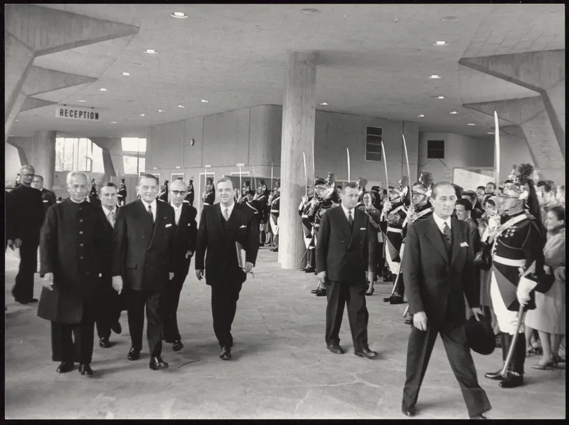 Photographie en noir et blanc : un cortège d'hommes en habit traverse un hall aux piliers de béton, entre deux haies de gardes républicains sabre au clair