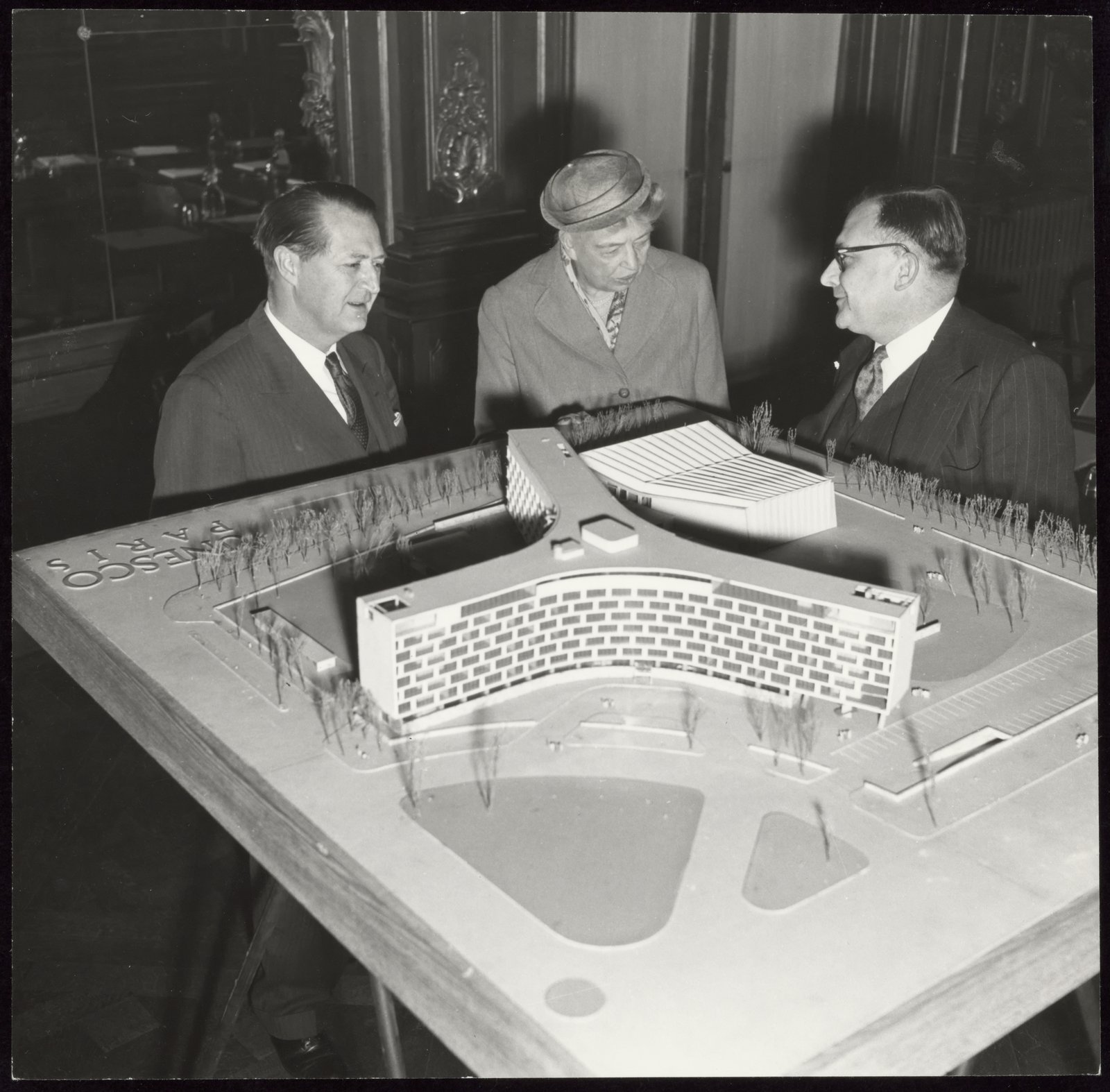 Eleanor Roosevelt examine la maquette du futur siège de la place de Fontenoy, 1955 — UNESCO · CC BY-SA 3.0 IGO — Archives de l’Unesco, Wikimedia Commons Photographie en noir et blanc : Eleanor Roosevelt, entre deux hommes en costume, se penche sur la maquette blanche du futur siège en étoile
