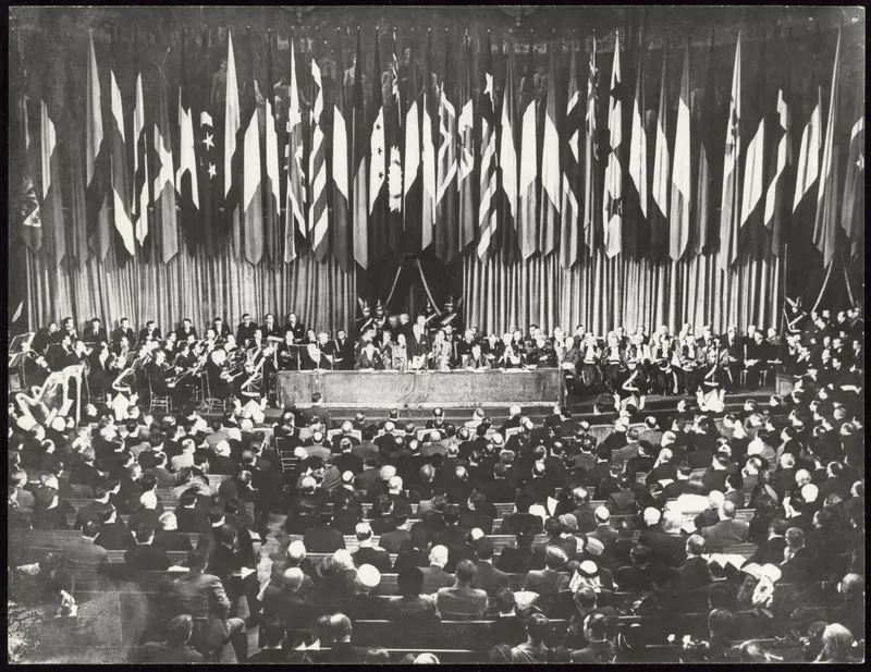 Photographie : un grand amphithéâtre plein, sous une rangée de drapeaux nationaux ; à la tribune, des orateurs, des musiciens et des professeurs en toge