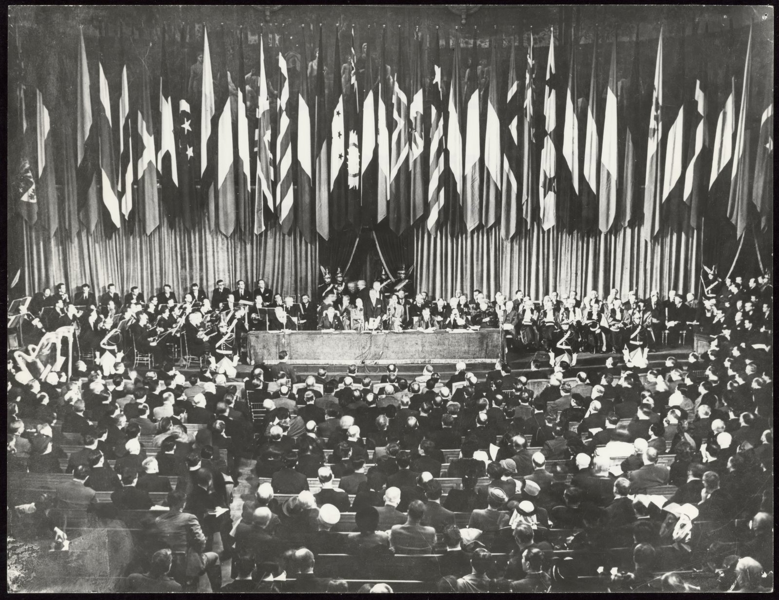 Ouverture de la première Conférence générale de l’Unesco à la Sorbonne, en novembre 1946 — UNESCO · CC BY-SA 3.0 IGO — Archives de l’Unesco, Wikimedia Commons Photographie : un grand amphithéâtre plein, sous une rangée de drapeaux nationaux ; à la tribune, des orateurs, des musiciens et des professeurs en toge
