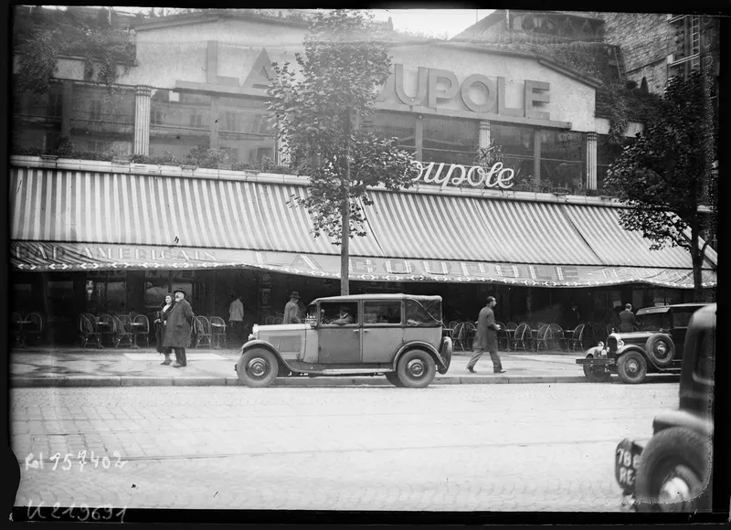 Photographie en noir et blanc : la terrasse de La Coupole sous ses stores rayés, l'enseigne « La Coupole » au-dessus, des automobiles des années 1930 garées devant