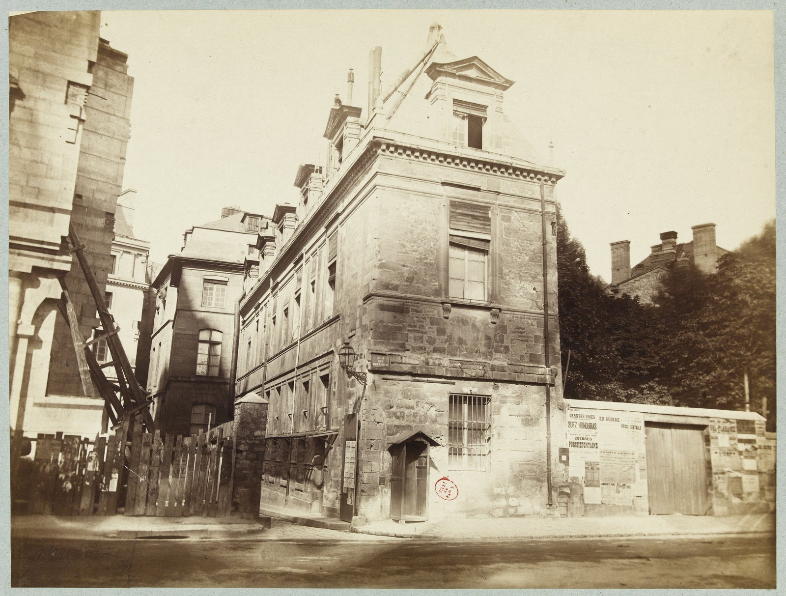 L’entrée de la rue de Jérusalem sur le quai des Orfèvres, photographiée par Pierre Emonts le 11 août 1869, avant sa disparition — Musée Carnavalet · CC0 — Paris Musées Photographie ancienne : à l'entrée d'une rue étroite, une maison de pierre à étage en angle, des palissades et des chantiers autour