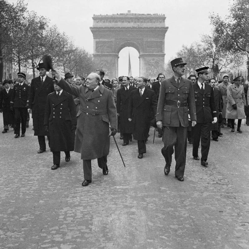 Photographie : Churchill, en manteau et casquette d'officier, et de Gaulle, en uniforme, marchent côte à côte sur les Champs-Élysées, suivis d'officiers, l'Arc de triomphe au fond