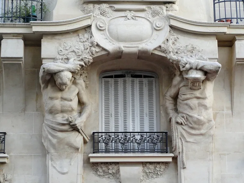 Photographie : une fenêtre encadrée de deux atlantes sculptés, sous un cartouche